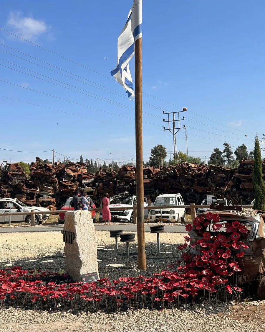 A memorial with an Israeli flag and red flowers in front of a pile of burnt cars