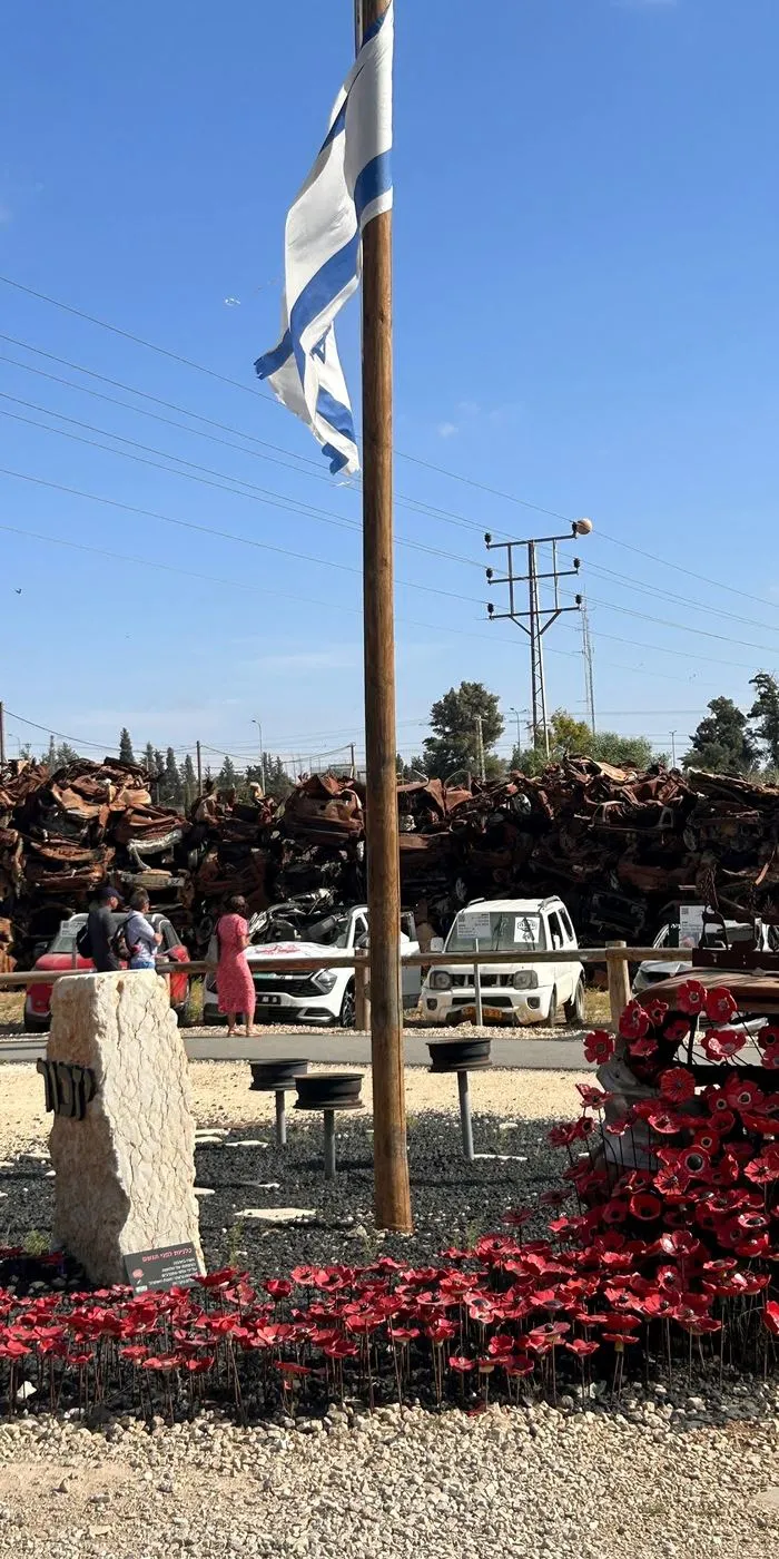 An Israeli flag over a memorial of red flowers, with burnt cars behind